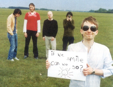 The five members of radiohead. Four of them are in the background; in the lower right corner you have a man holding a sign that reads 'if we smile, can we go?'.